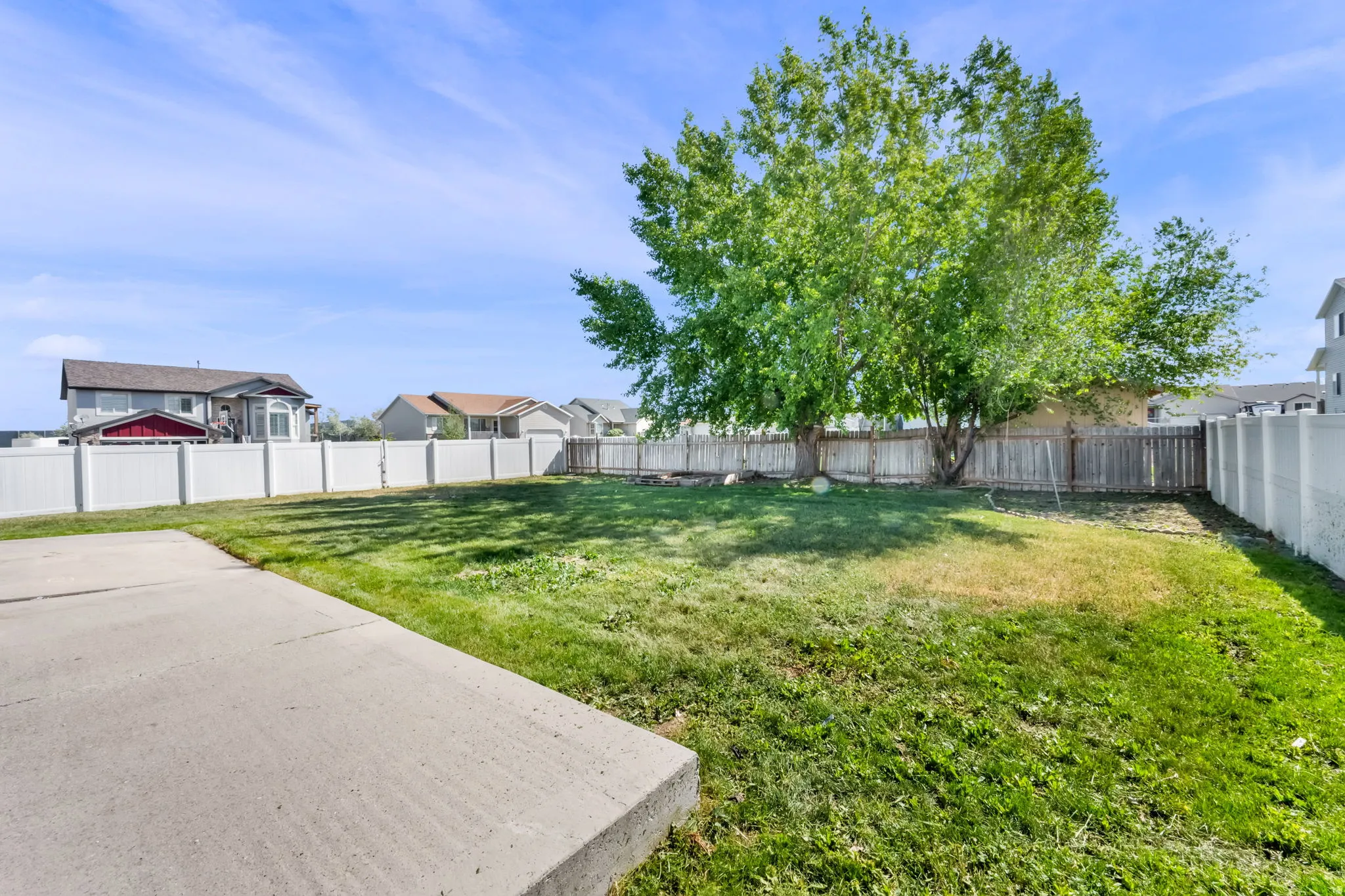 Spacious backyard featuring a large concrete patio, mature trees, and a combination of white vinyl and wood fencing