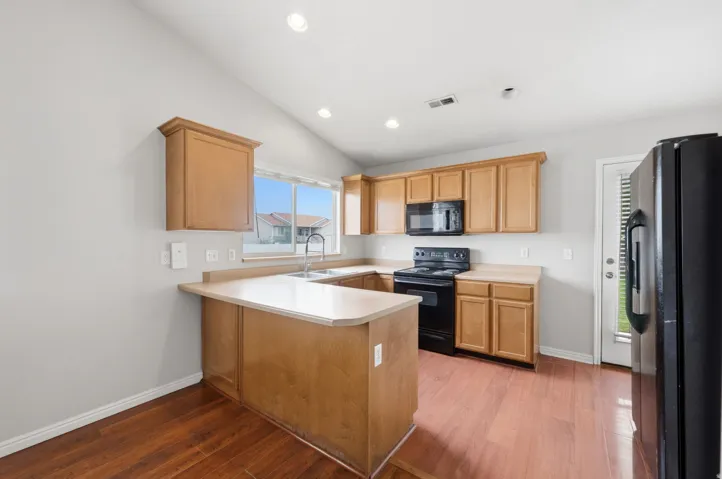Kitchen featuring wood-finish flooring, light wood cabinetry, light-toned countertops, a stainless steel sink, and black appliances