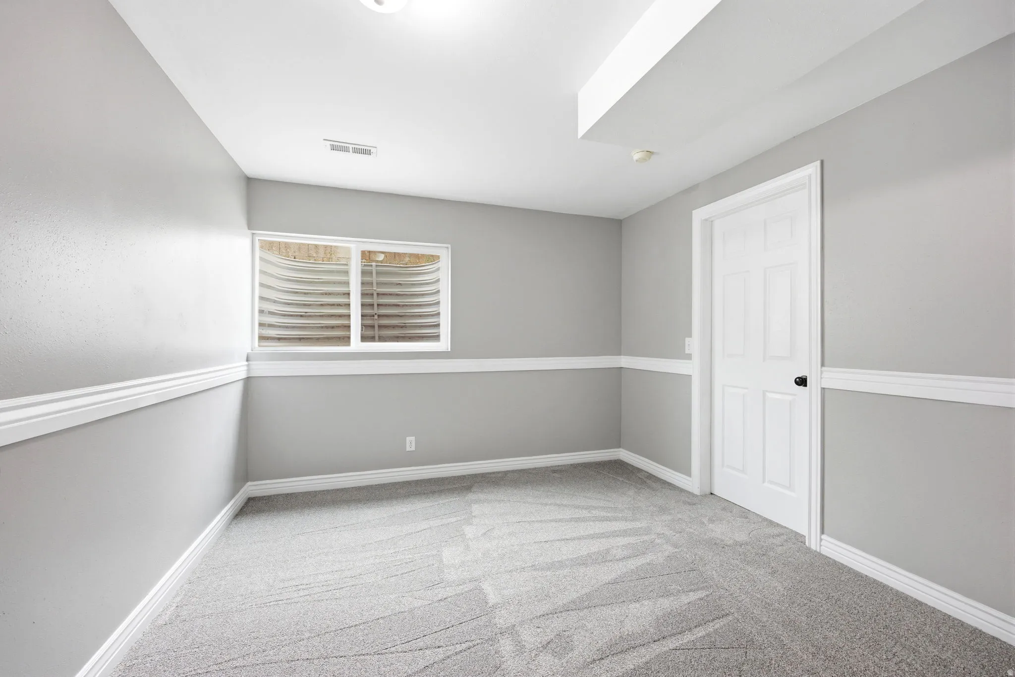 Carpeted room featuring gray walls with white chair rail molding, a white paneled door with black hardware, and a window with a window well