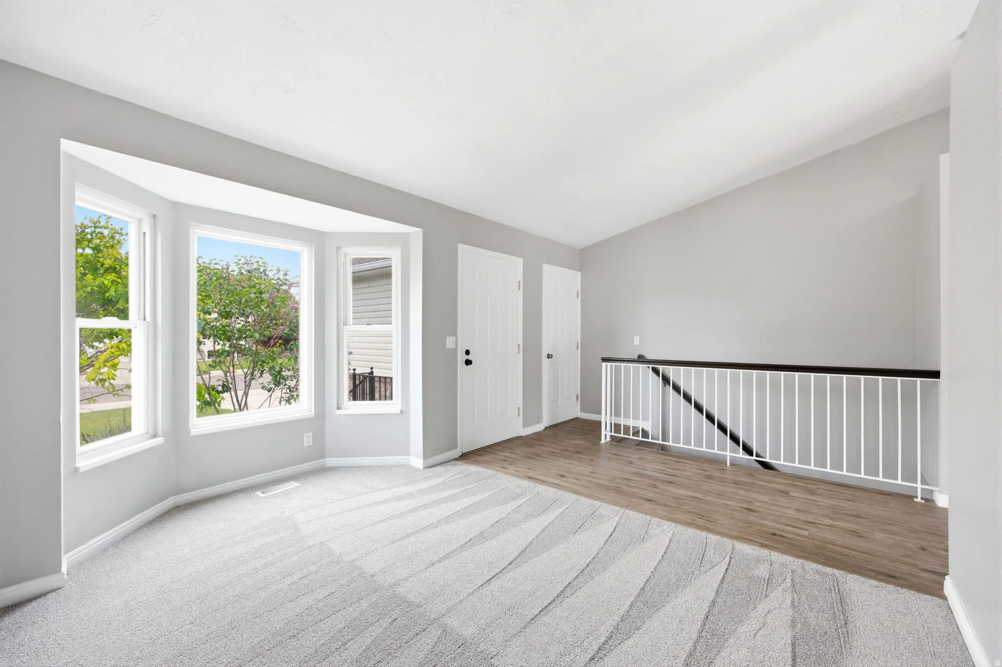 Bay window with multiple vertical windows, light gray walls, textured gray carpeting, wood-finish flooring, and a white baluster railing with a dark top rail