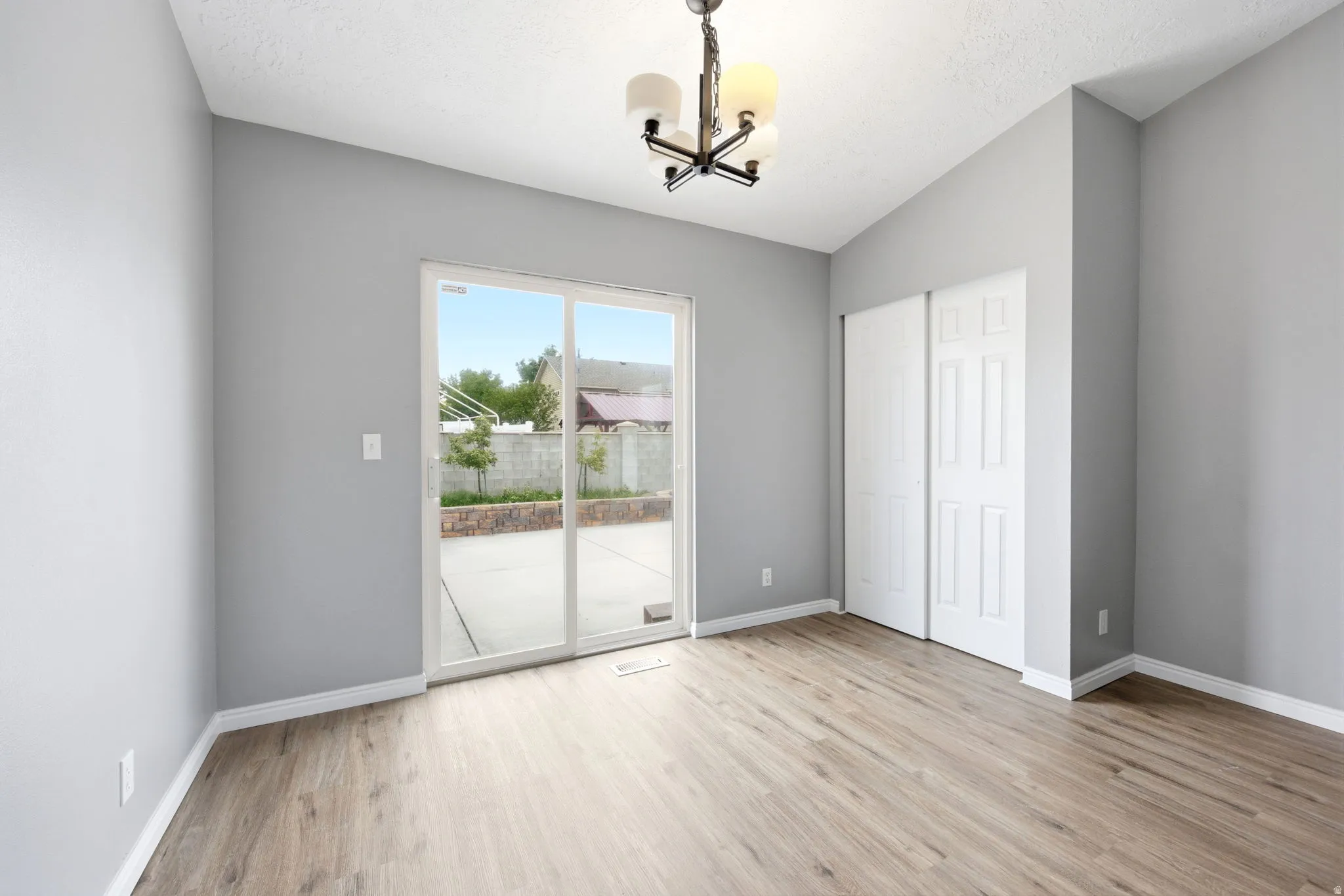 Open room featuring wood-finish flooring, light gray wall finishes, a vaulted ceiling, a black and white chandelier, and a sliding glass door providing access to a concrete patio