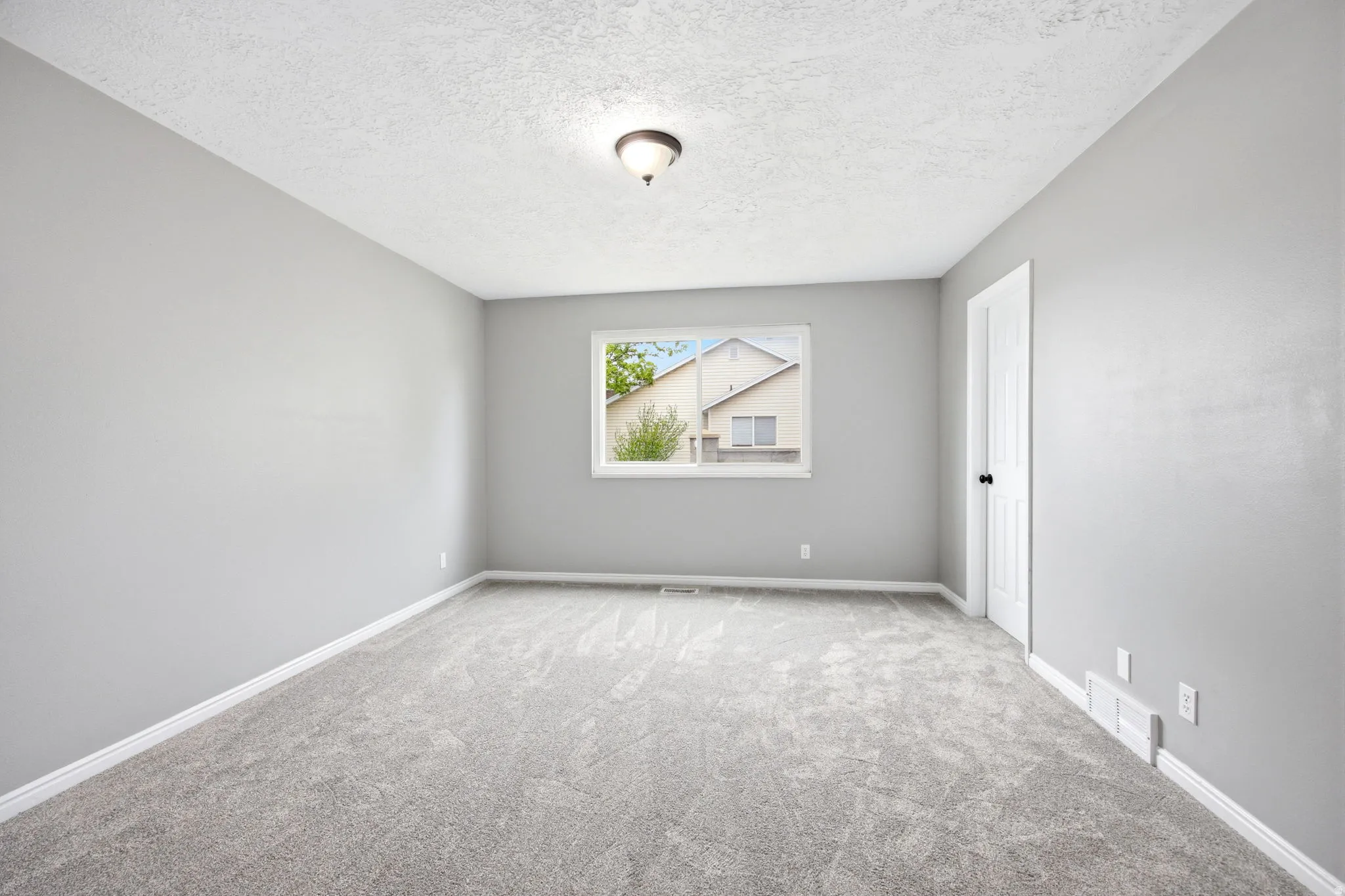 Spacious room featuring light grey walls, grey carpet flooring, a white window frame, white baseboards, and a ceiling-mounted light fixture