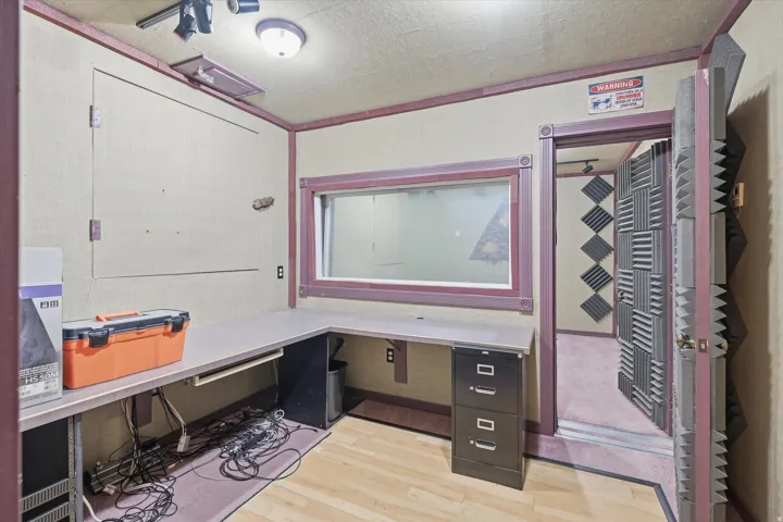 Dedicated workspace featuring a built-in L-shaped desk, a window connecting to an interior room, and a door with acoustic paneling