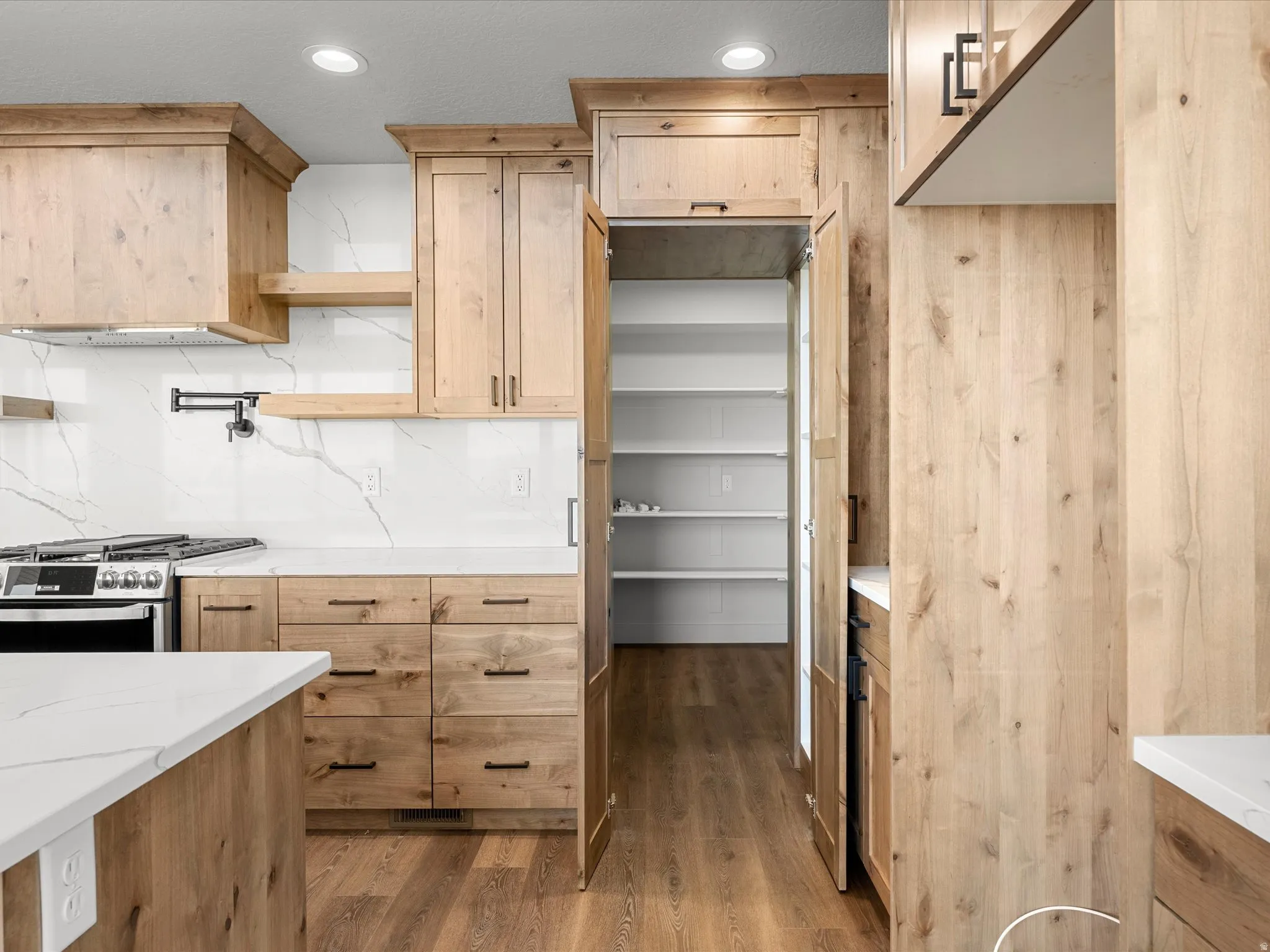 Kitchen featuring wood-finish cabinetry, white marble-look countertops and backsplash, stainless steel range with pot filler, and a walk-in pantry with ample shelving