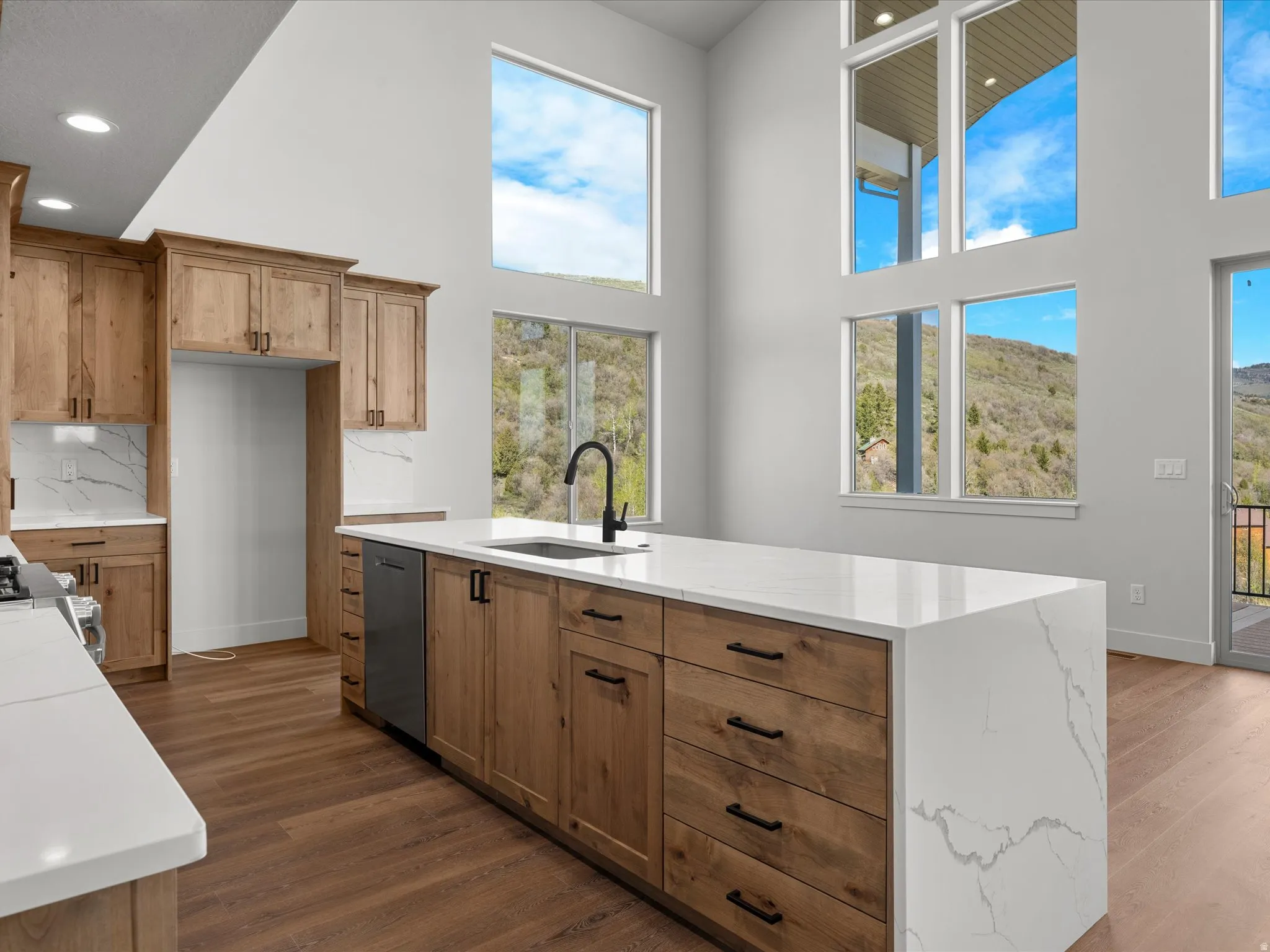 Open-concept kitchen featuring extensive wood cabinetry, a large waterfall island with a sink and a black faucet, white countertops with veining, and wood-finish flooring