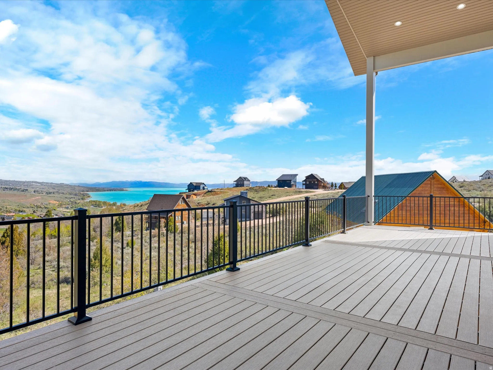 Expansive outdoor decking with composite wood-finish flooring, featuring a black metal railing system, covered patio area with recessed lighting, and a panoramic lake and mountain backdrop