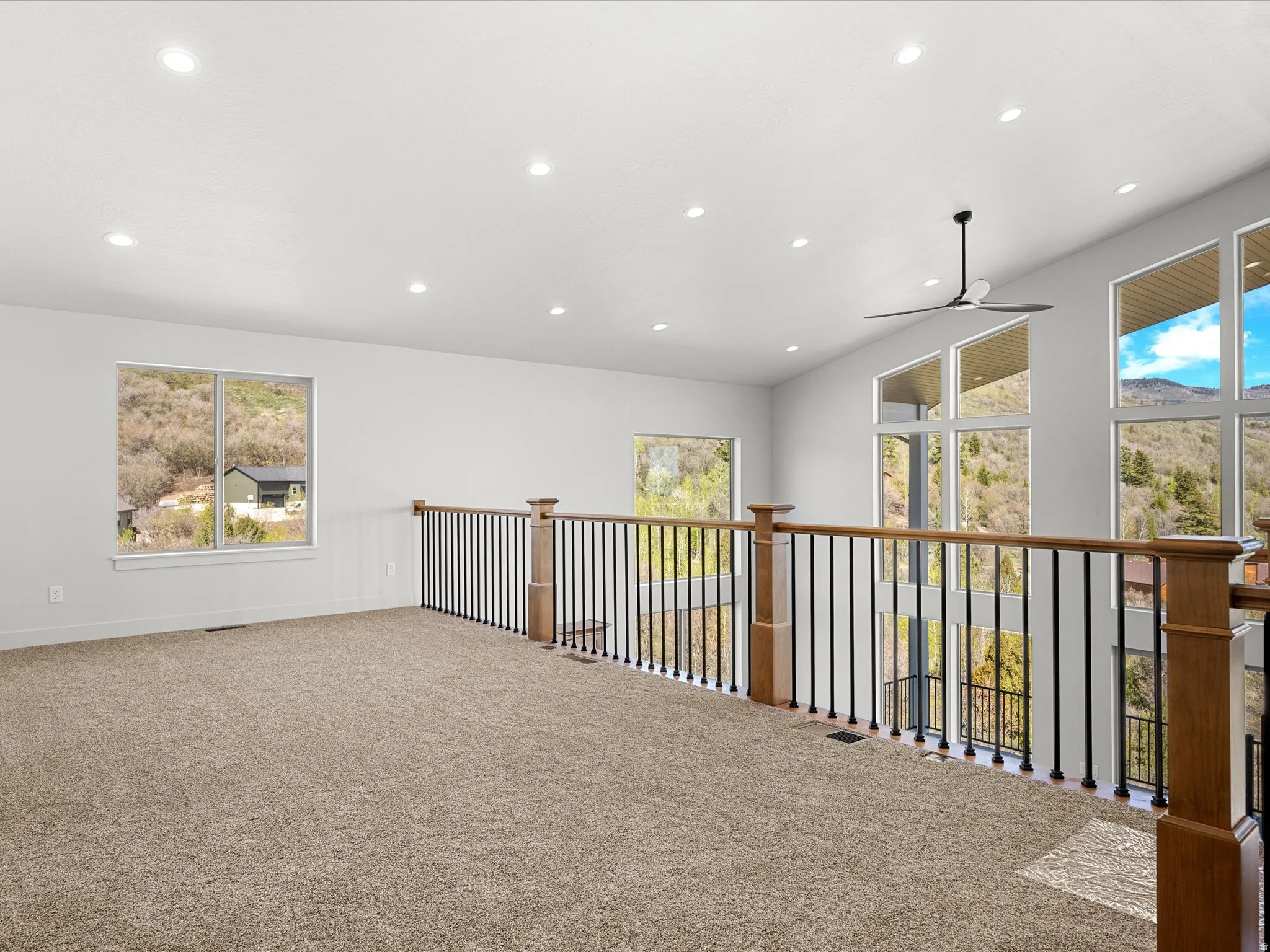 Expansive loft space with neutral carpeting, recessed ceiling lighting, a contemporary ceiling fan, a wood-finish and wrought iron railing, and large windows with mountain scenery