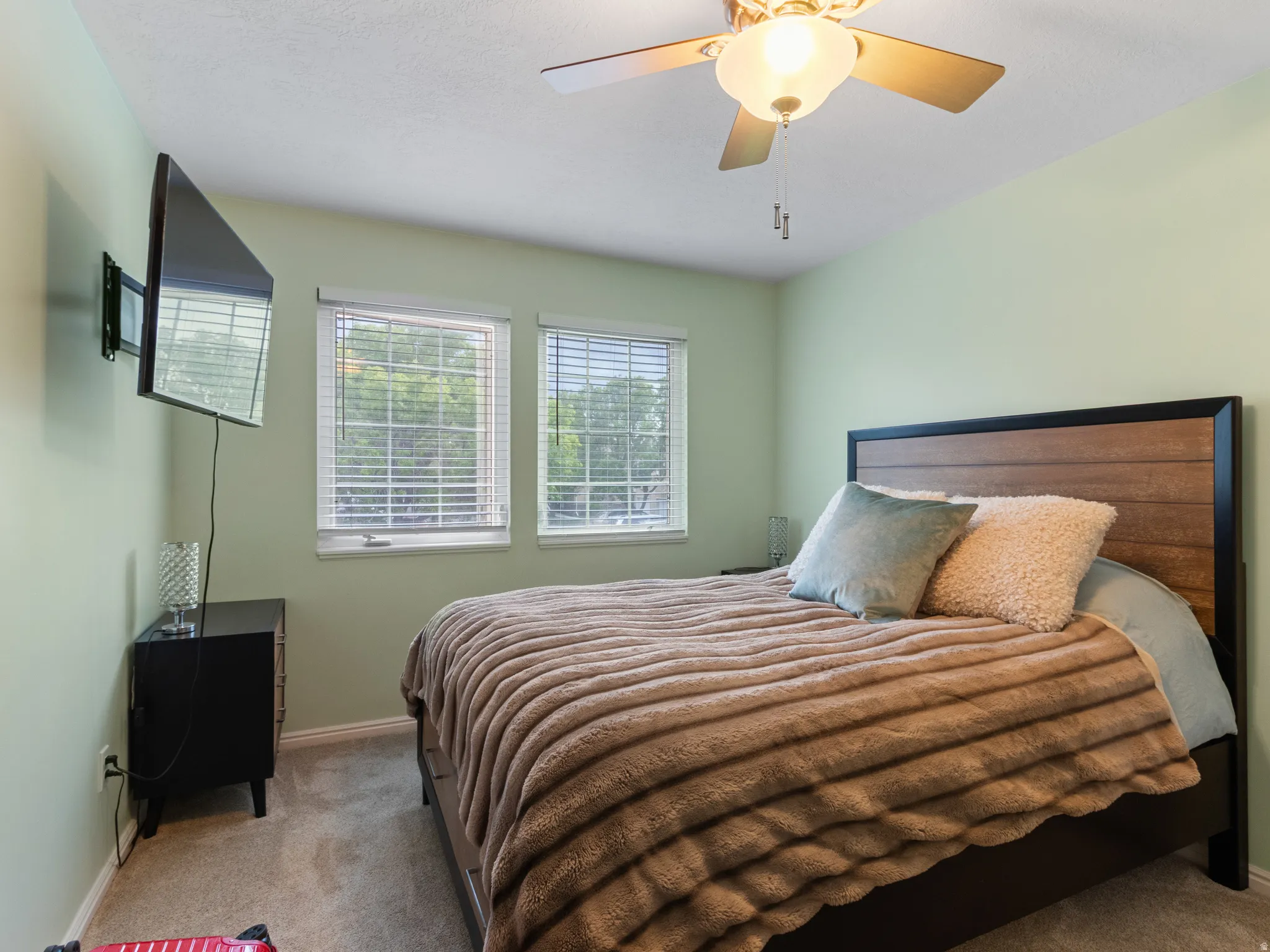 Carpeted bedroom featuring two windows with horizontal blinds, a wood-finish headboard, and a ceiling fan with light fixture