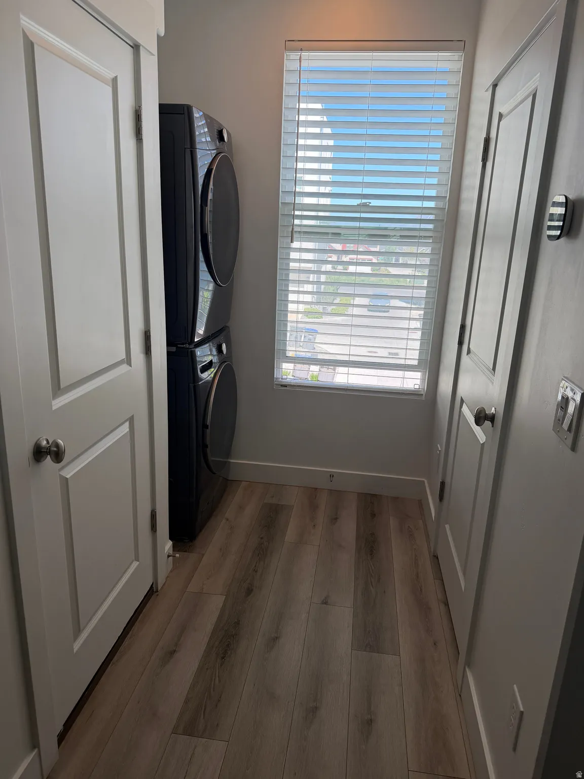 Dedicated laundry area featuring wood-finish flooring, a stacked washer and dryer unit, and a window with horizontal blinds