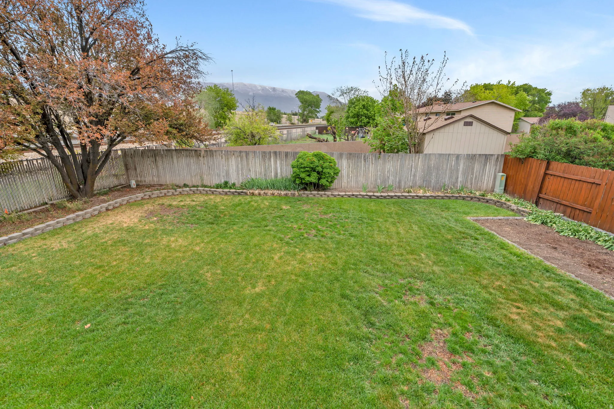 Expansive backyard featuring a manicured lawn, a mature tree, a privacy fence, a retaining wall, and a dedicated garden bed