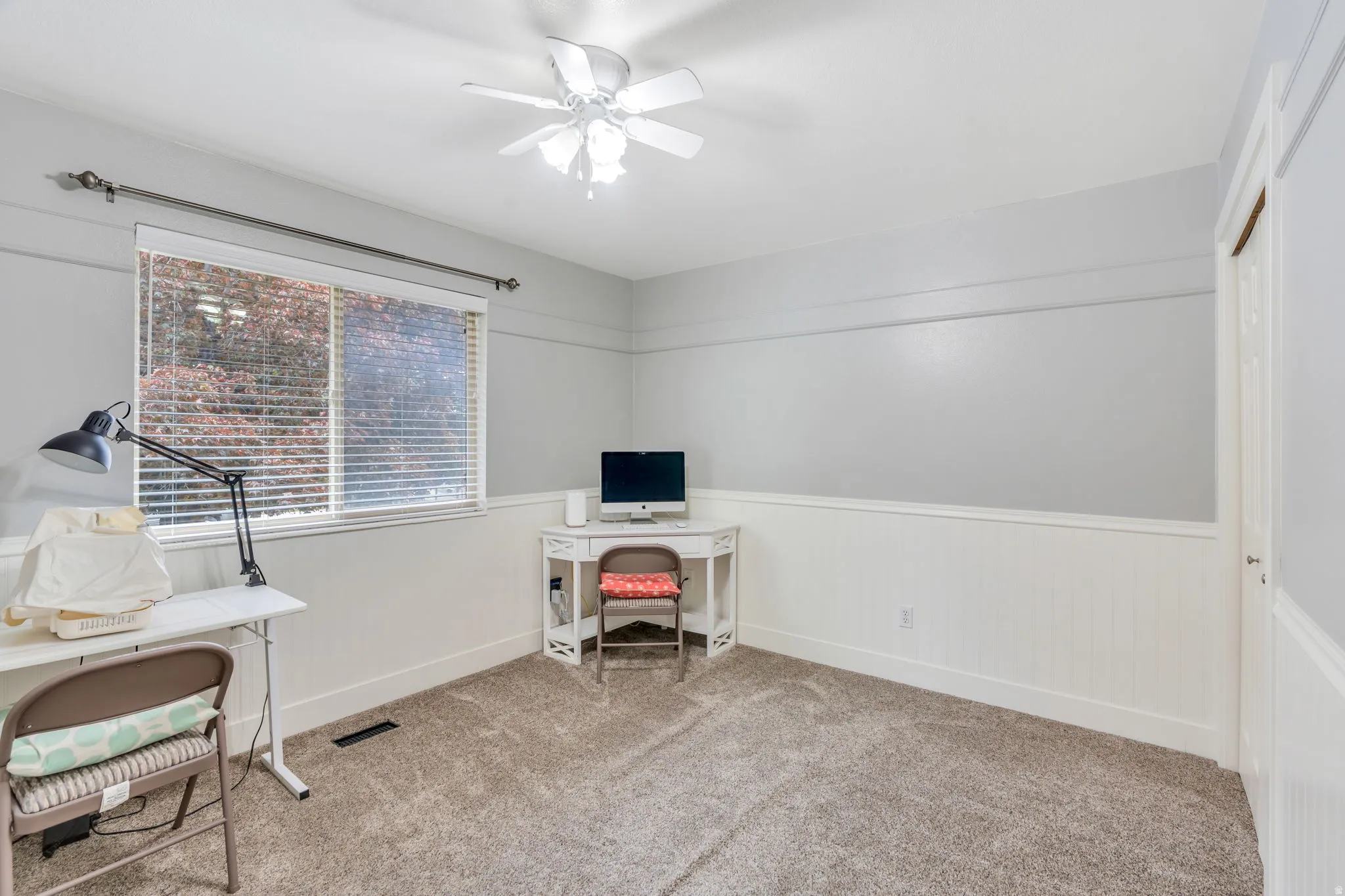 Carpeted room featuring a large window with horizontal blinds, a white ceiling fan, and white wainscoting