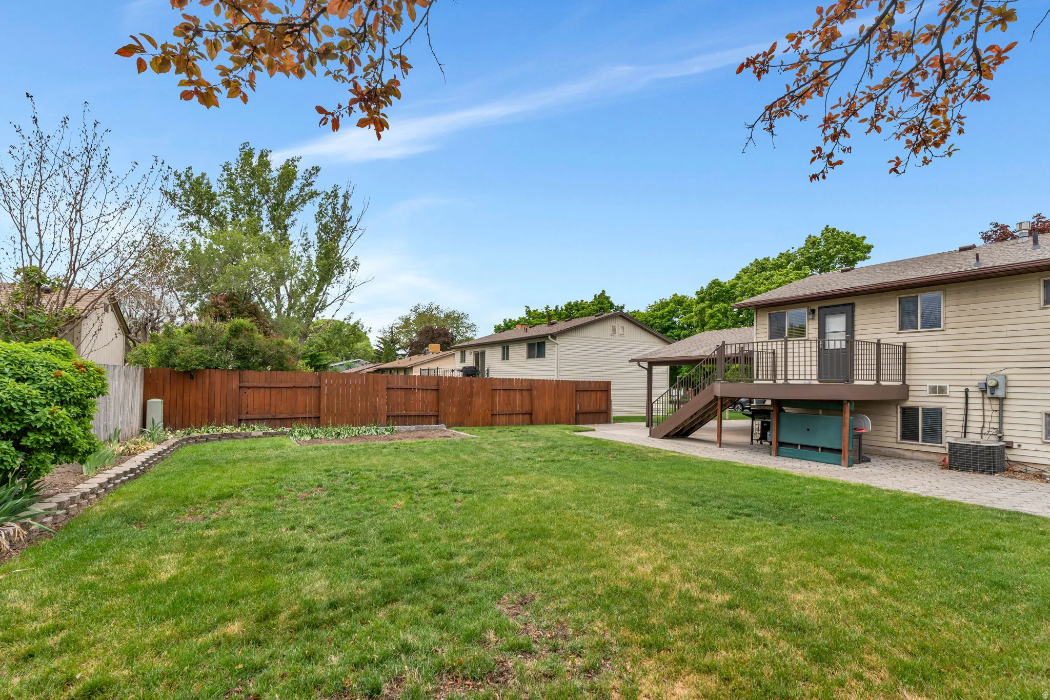 Expansive backyard featuring a manicured lawn, a raised wood deck with metal railings, a ground-level paver patio, a full-height privacy fence, and an integrated garden bed