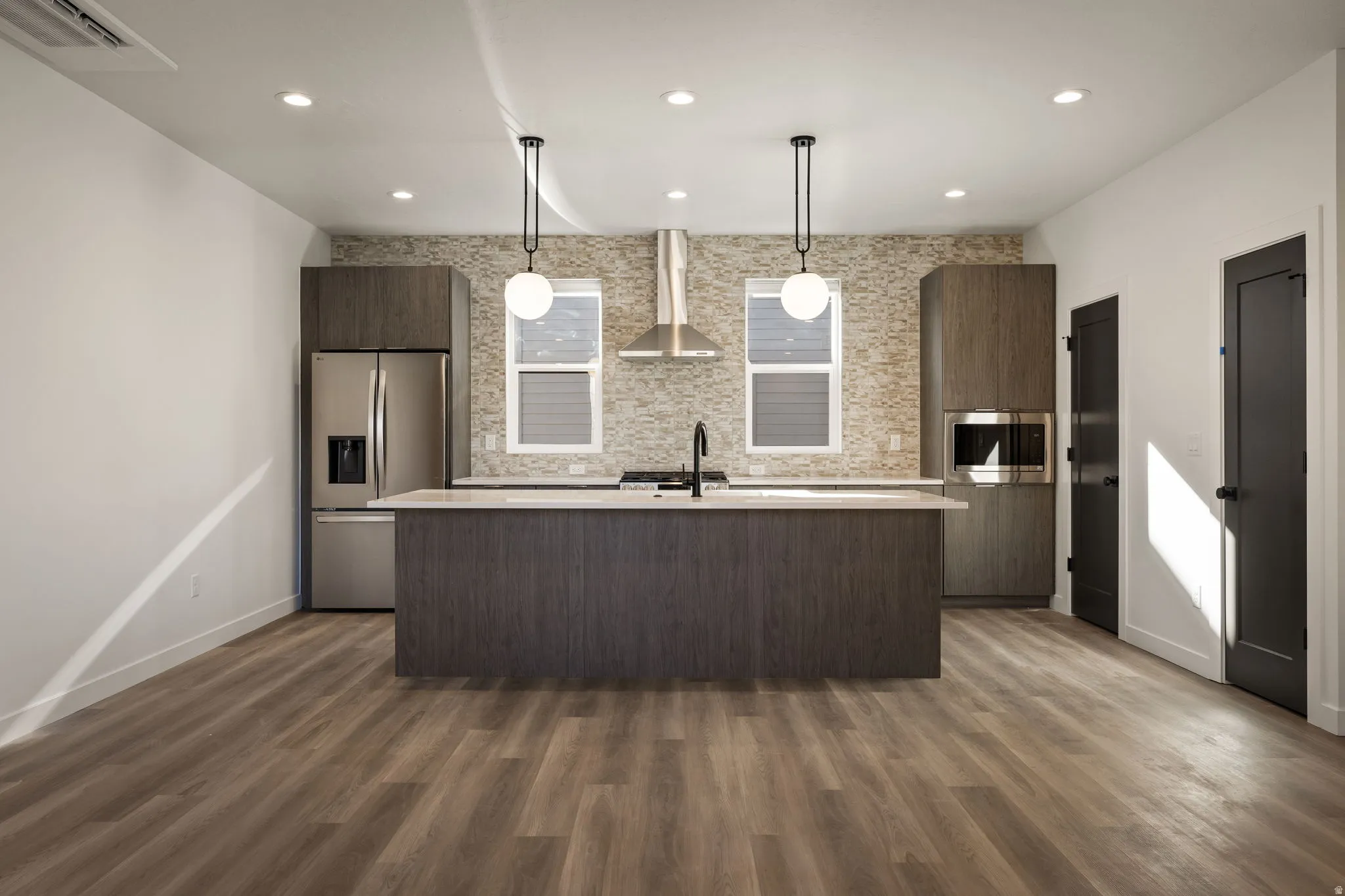 Modern kitchen featuring a central island with a light-toned countertop, stainless steel appliances, wood-finish flooring, recessed ceiling lighting, and pendant fixtures
