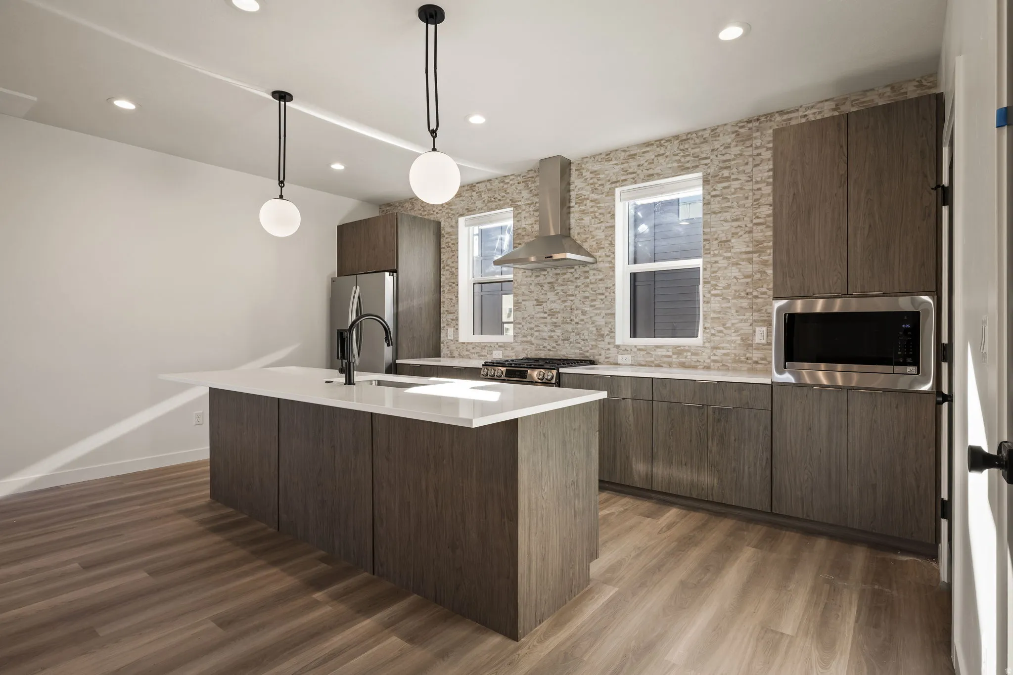 Modern kitchen featuring an island with a built-in sink and white countertop, wood-finish cabinetry, stainless steel appliances, and a textured stone-look backsplash