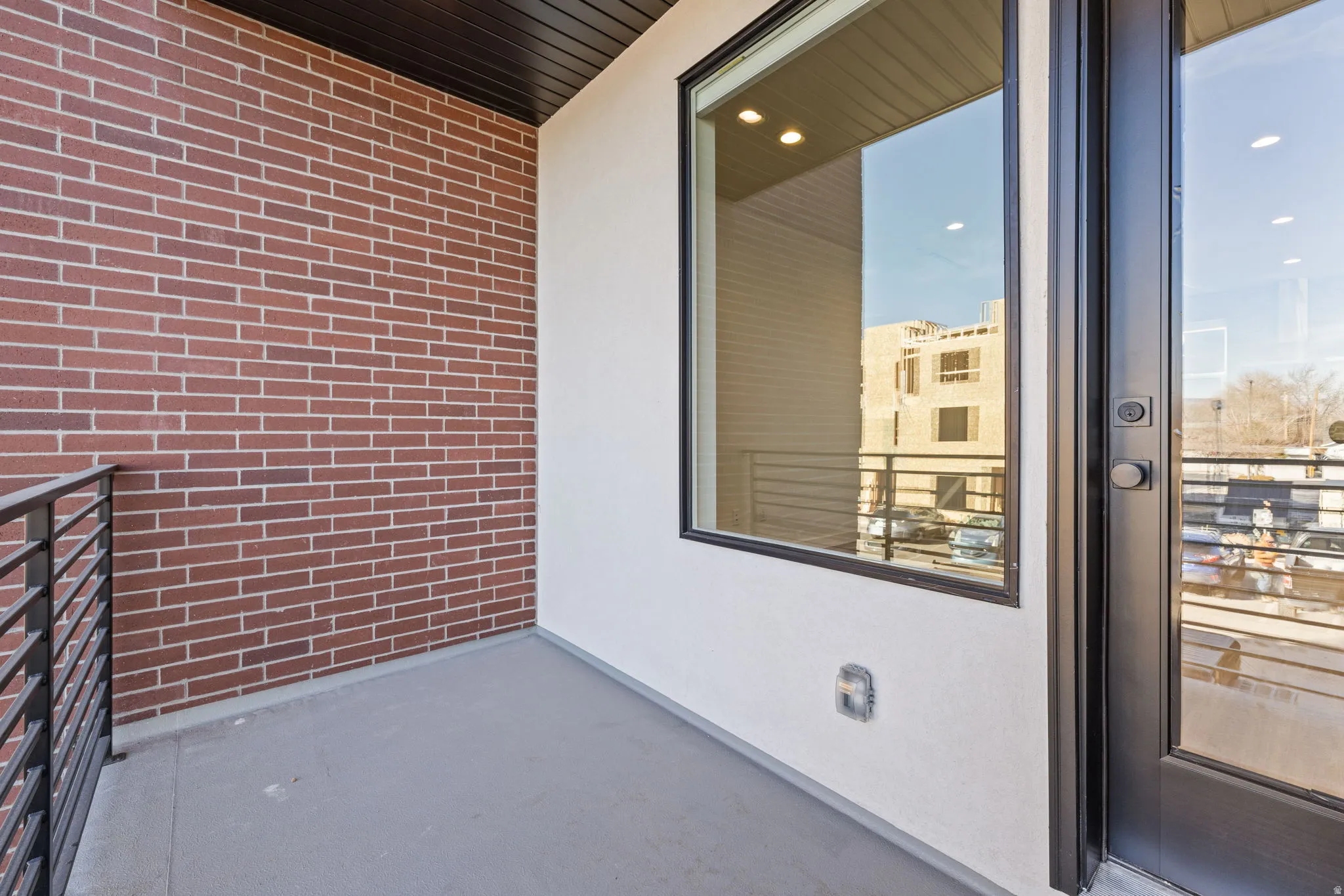 Outdoor balcony featuring a brick accent wall, a large fixed window, a glass-paneled door, metal railing, and a dark wood-finish soffit