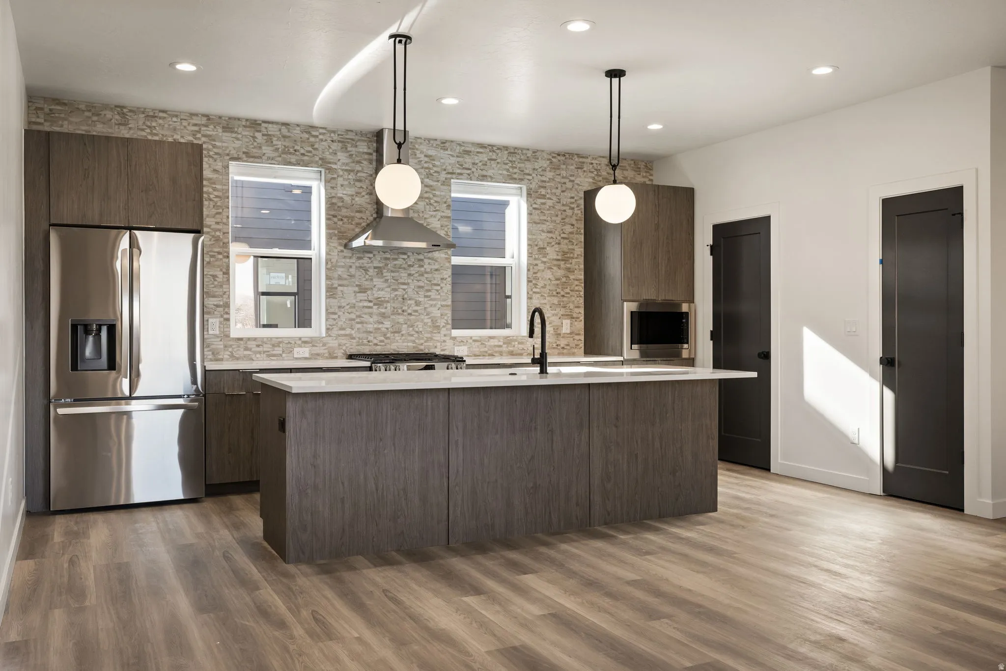 Modern kitchen featuring wood-finish flooring, a central island with a built-in sink, stainless steel appliances, and sleek dark cabinetry