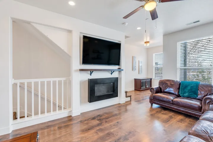 Spacious living area featuring wood-finish flooring, a wall-mounted fireplace with a mantel shelf, and a ceiling fan with integrated lighting