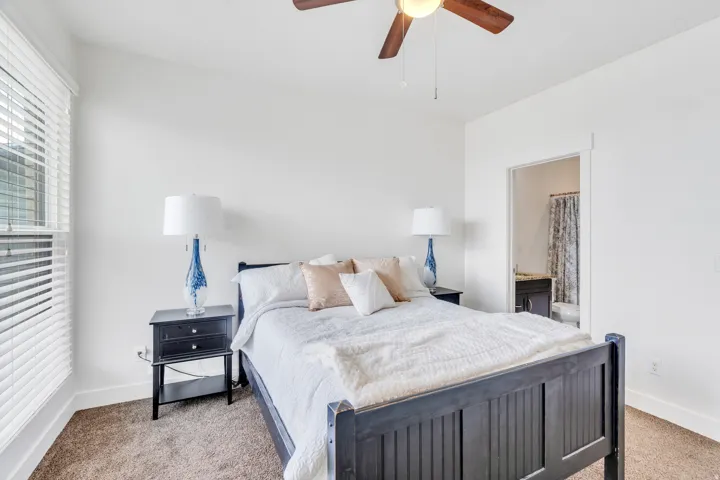 Carpeted bedroom with a ceiling fan, a window featuring horizontal blinds, and a doorway revealing a bathroom with a vanity and shower