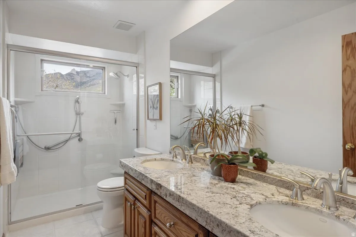 Spacious bathroom featuring a dual vanity with a speckled countertop, a large wall-mounted mirror, white floor tiling, a walk-in shower with glass enclosure, and a frosted window showcasing mountain scenery
