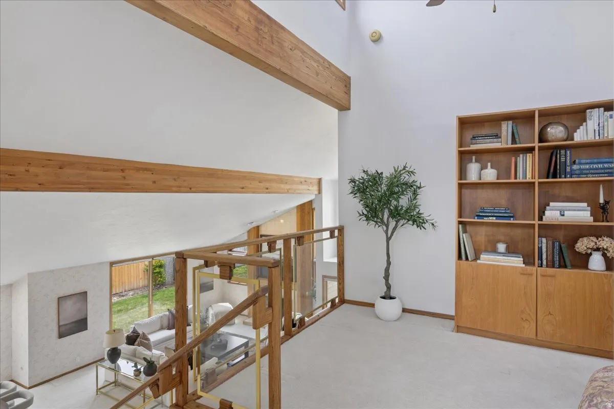 Loft area featuring a vaulted ceiling with exposed wood beams, a built-in wooden bookcase, and a wooden handrail with glass panels overlooking the lower level