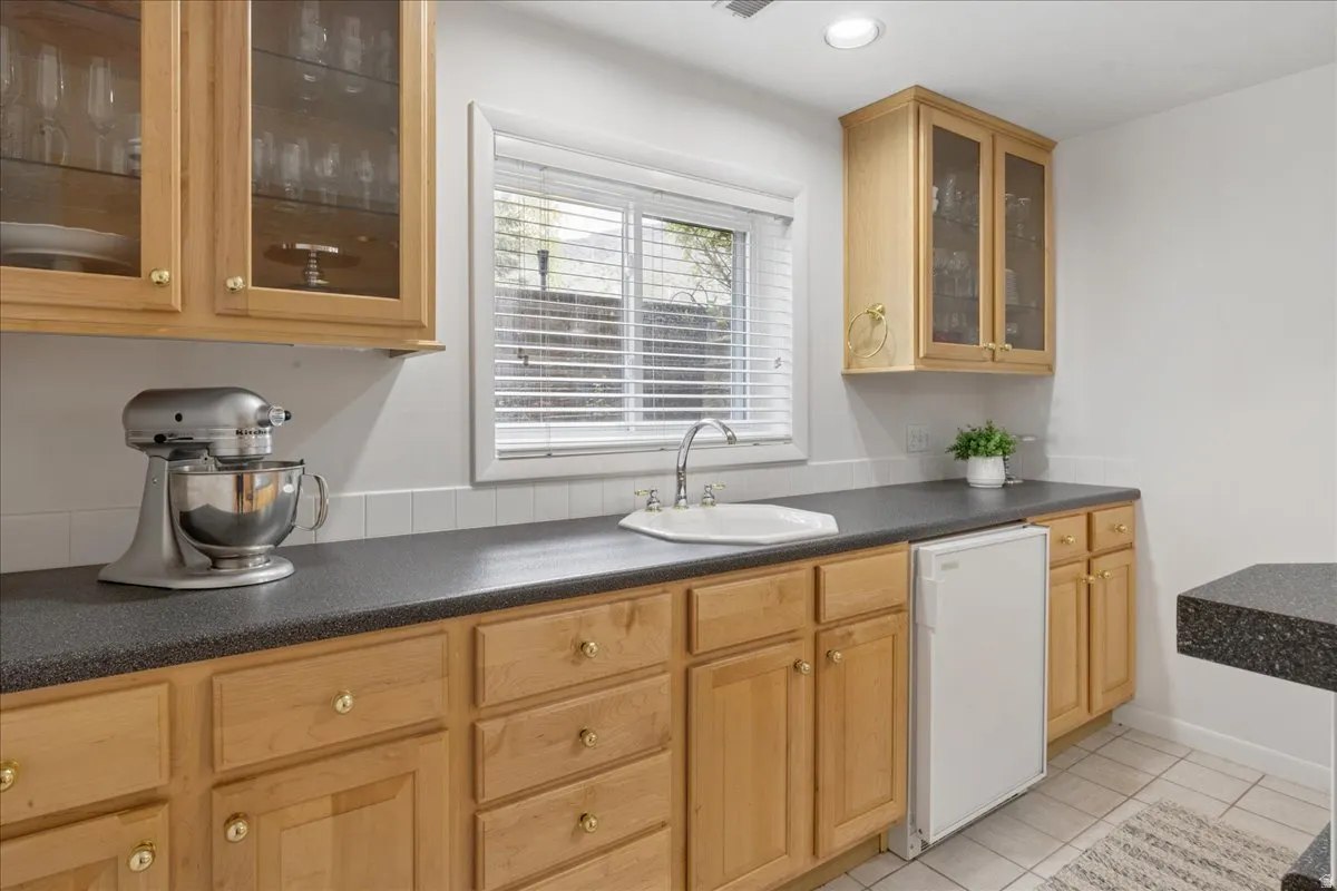 Kitchenette featuring blonde wood cabinetry with brass hardware, dark countertops, a white rectangular basin sink with a chrome faucet, and a built-in compact refrigerator