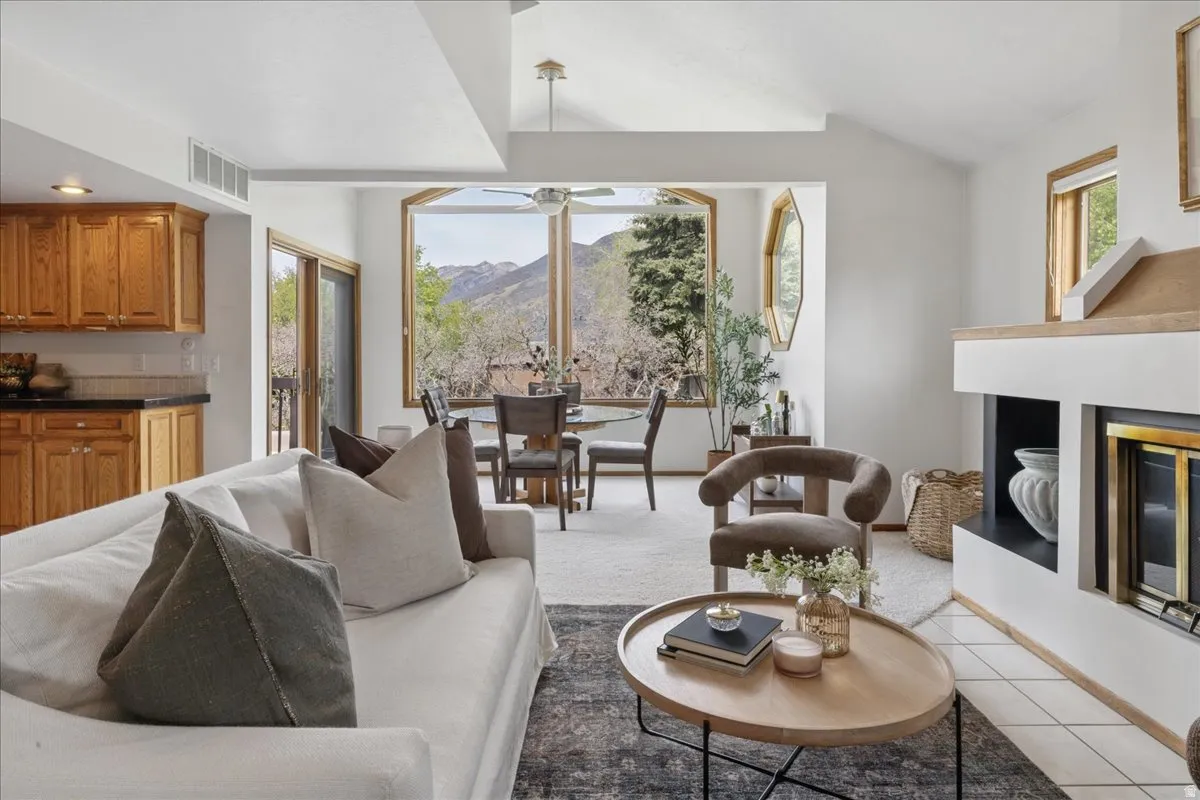 Bright living space with vaulted ceilings and large windows, featuring a built-in fireplace, tile flooring, and a kitchen area with wood cabinetry and dark countertops