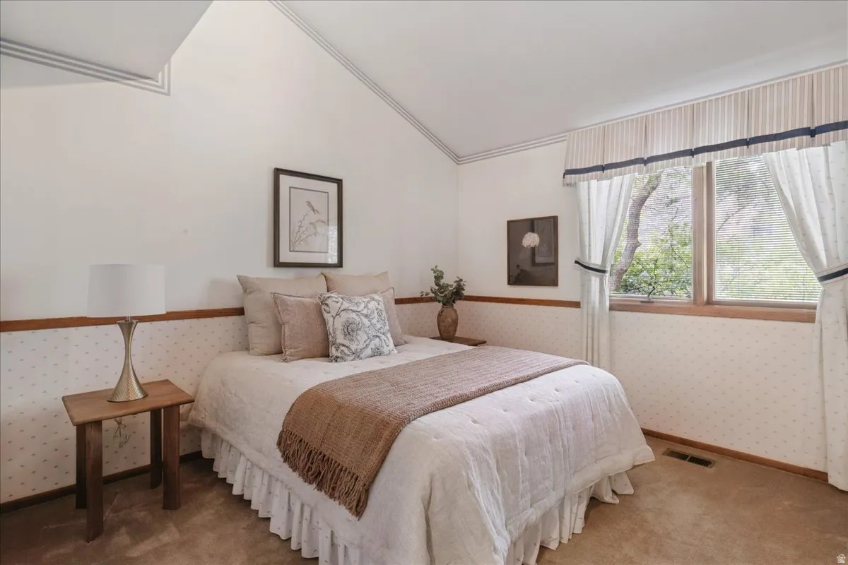 Vaulted ceiling with angled architectural details, wood-trimmed window, patterned wall covering, carpeted flooring, and a built-in wall picture frame