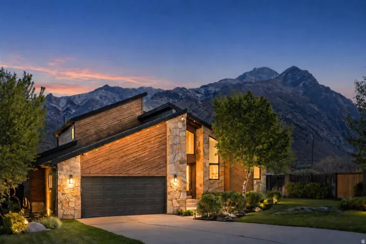 Modern architectural residence featuring a stone and wood-paneled exterior, an integrated two-car garage, and landscaped front yard with established plantings