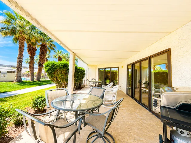 Expansive covered patio featuring a tiled floor, stucco exterior, multiple sets of glass sliding doors, and surrounding landscaping with palm trees