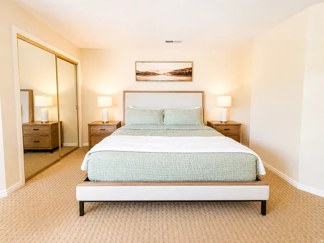 Wall-to-wall patterned carpeting, mirrored closet doors, a bed frame with a wood-finish and upholstered headboard, cream-toned walls, and white baseboards
