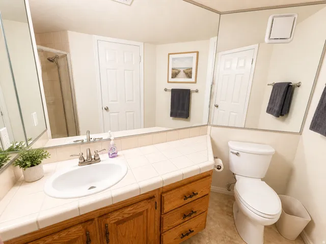 Bathroom vanity featuring a light wood-finish cabinet with brass-tone hardware, a tiled countertop, and an oval undermount sink with a chrome faucet