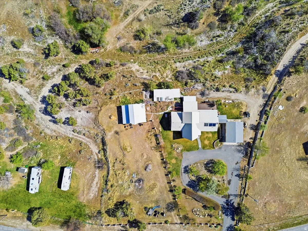 Aerial perspective showcasing a sprawling property featuring a main residence with a light-colored roof, multiple outbuildings, extensive natural landscaping, a paved driveway, and a circular turnaround area