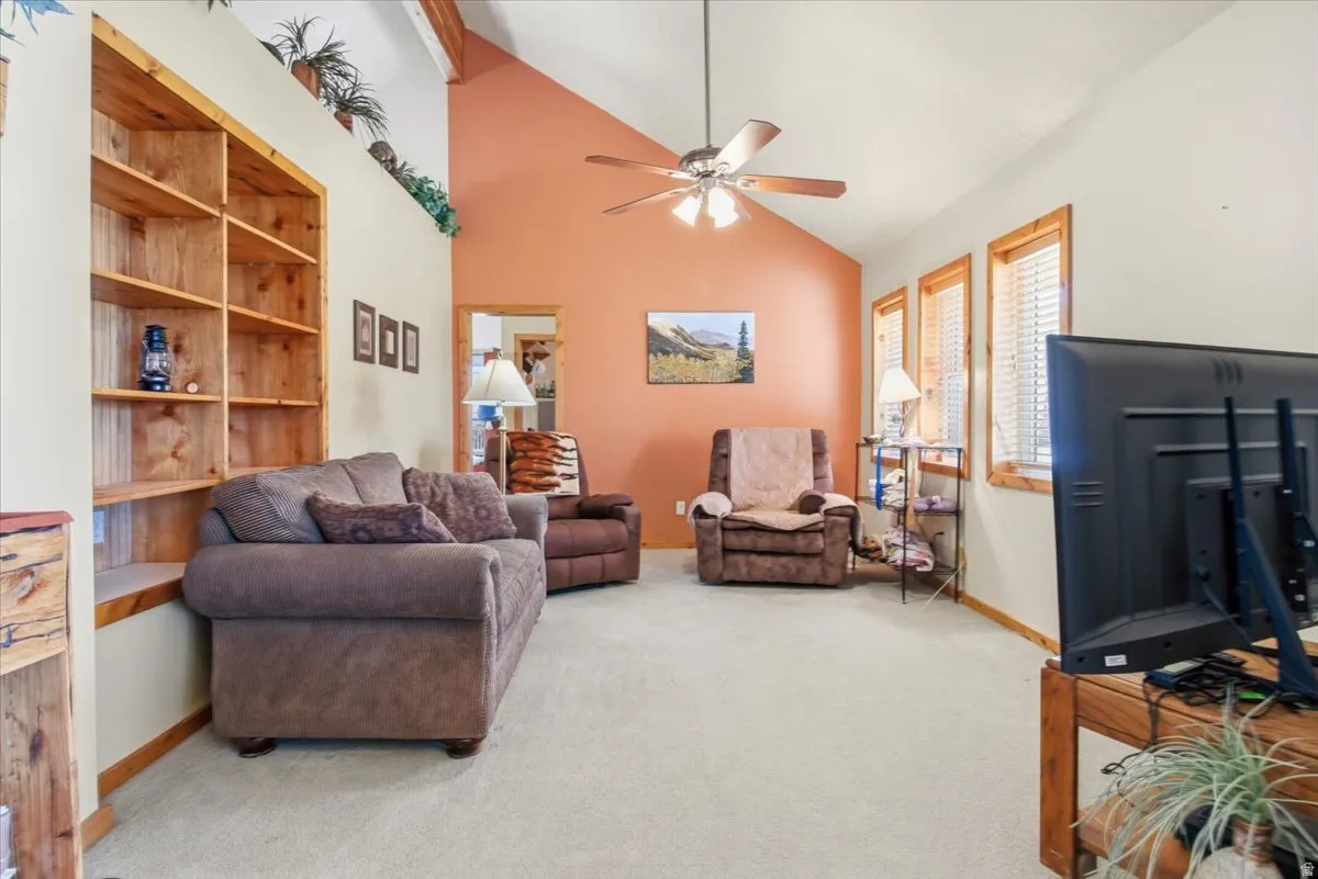 Spacious living area featuring a vaulted ceiling with exposed wood beams, a ceiling fan, and light beige carpeting