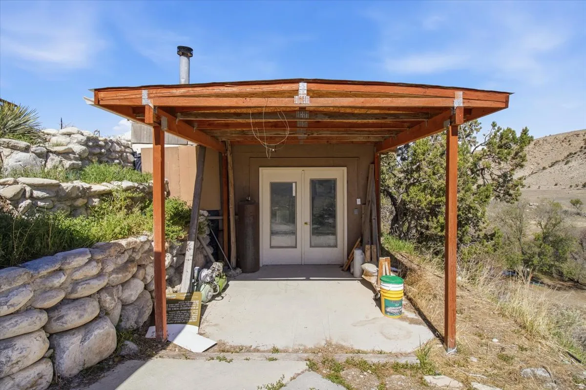 Covered concrete patio featuring a natural wood pergola, double exterior doors, a stacked stone retaining wall, and natural landscaping with mature trees