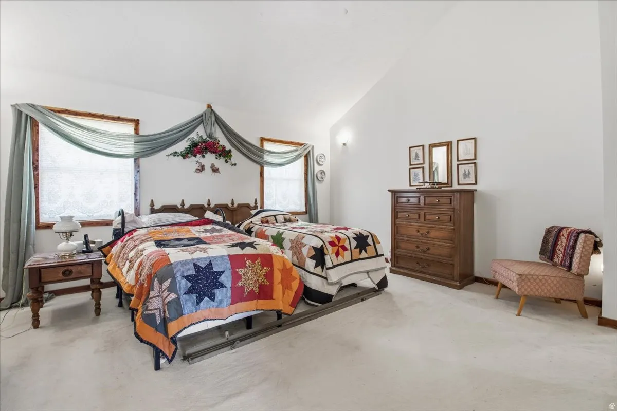 Room featuring a vaulted ceiling, two windows with wood trim, wall-mounted sconces, and light-colored carpeting