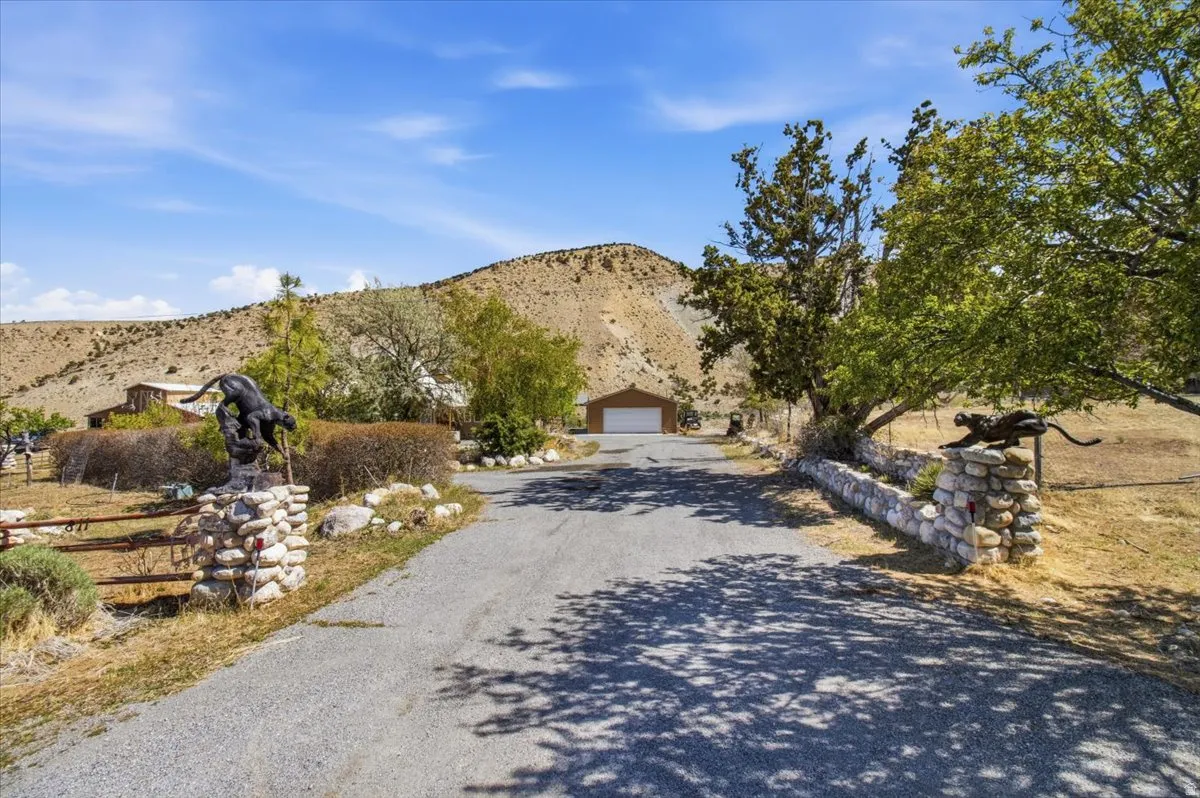 Gravel driveway entrance featuring stone pillars with decorative animal sculptures, mature trees, and a distant two-car garage with a brown exterior