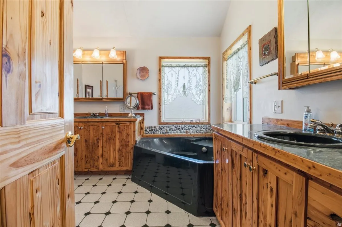 Bathroom featuring a corner soaking tub, dual wood-finish vanities with under-mount sinks, wood-finish framed mirrors with integrated lighting, and patterned tile flooring