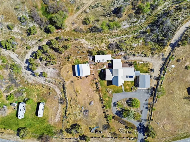 Aerial perspective showcasing a sprawling property featuring a main residence with a light-colored roof, multiple outbuildings, extensive natural landscaping, a paved driveway, and a circular turnaround area