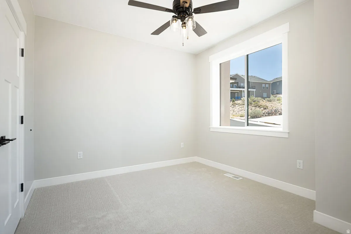 Neutral-toned room featuring a window with a white frame and trim, grey carpet flooring, light grey wall paint, and a dark-toned ceiling fan with integrated lighting