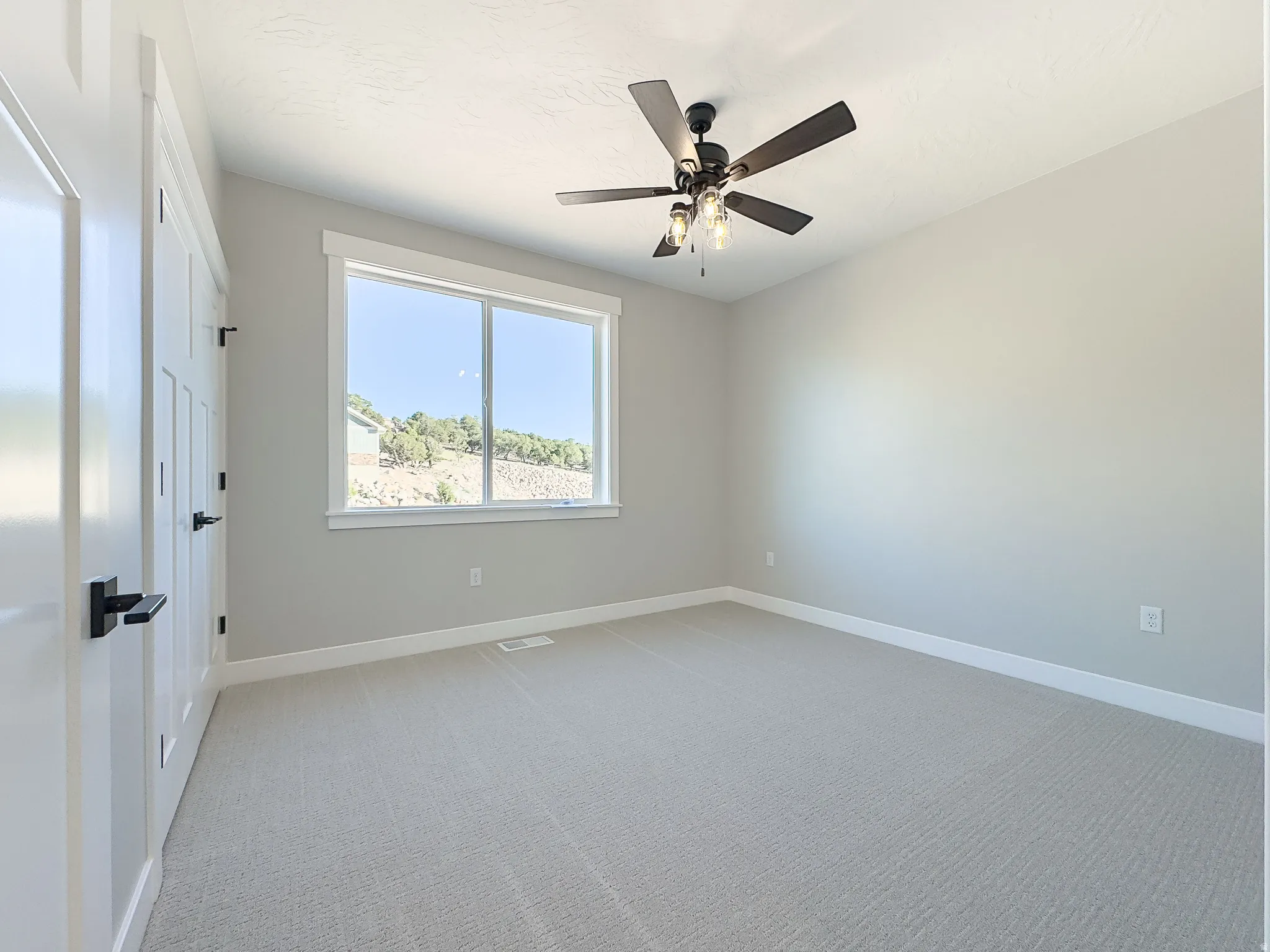 Spacious room featuring neutral carpeting, light gray walls, a ceiling fan with integrated lighting, and a large window with white trim