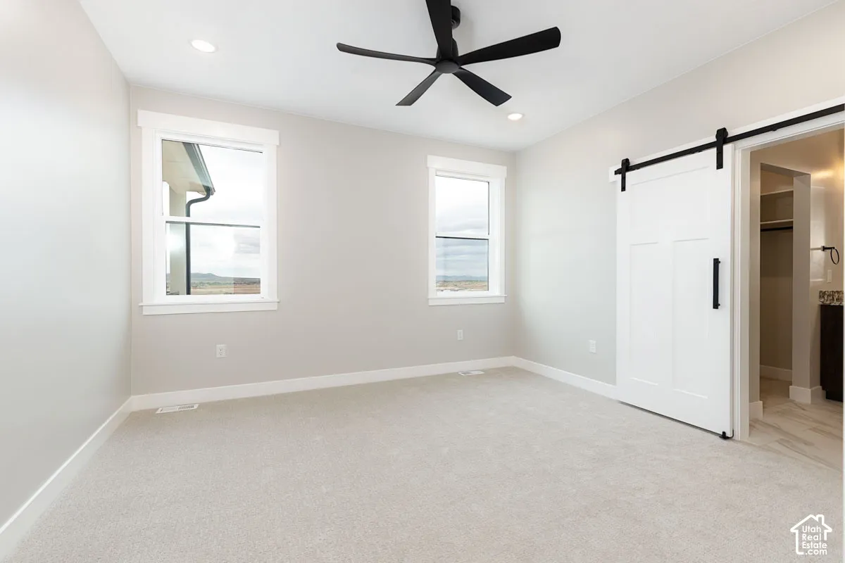 Bright room with light gray carpeting, two large windows with white trim, a black ceiling fan, and a white barn door with black hardware