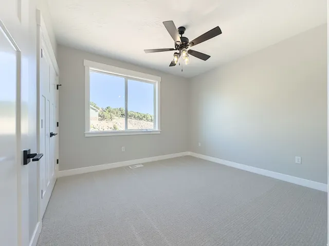 Spacious room featuring neutral carpeting, light gray walls, a ceiling fan with integrated lighting, and a large window with white trim