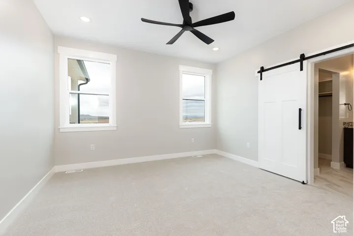 Bright room with light gray carpeting, two large windows with white trim, a black ceiling fan, and a white barn door with black hardware