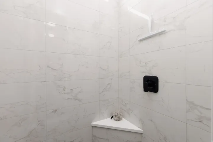 Shower enclosure featuring a white tile surround with subtle gray veining, a matte black shower control, a white shower head, and an integrated corner shelf