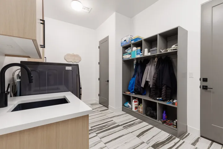 Utility room featuring a white countertop with integrated sink and matte black faucet, custom wood-finish cabinetry, built-in gray storage cubbies and coat hooks, and striped tile flooring