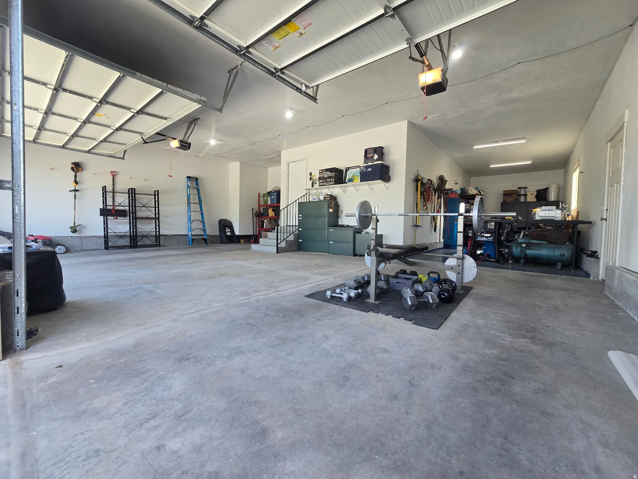 Spacious garage featuring concrete flooring, white walls, multiple garage door openers, built-in shelving, and a set of interior stairs with a black metal railing
