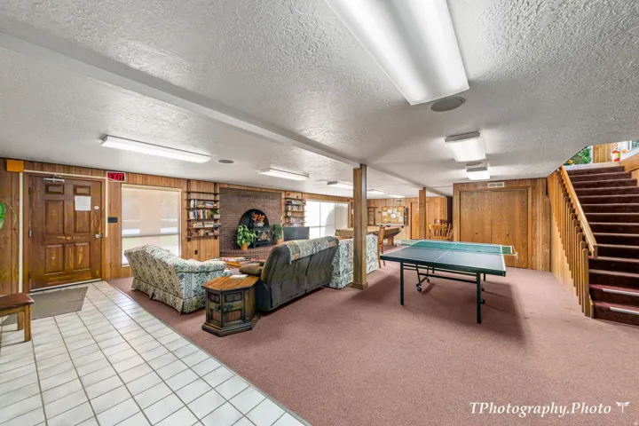 Spacious recreation room featuring wood paneling, a brick fireplace, built-in shelving, carpet flooring, and an interior staircase with wood railings