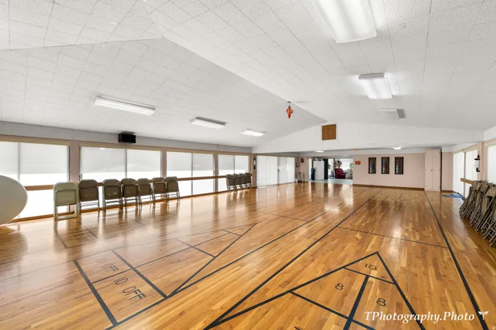 Spacious open-concept room featuring wood-finish flooring with shuffleboard court markings, numerous windows with roller shades, an acoustic panel ceiling, and recessed lighting