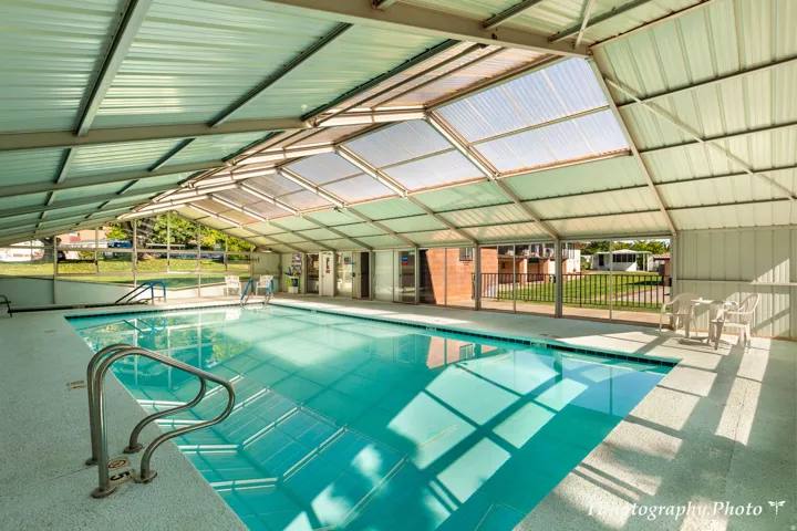 Indoor swimming pool with clear water, featuring a metal roof structure, translucent skylight panels, and an open side wall with horizontal fencing