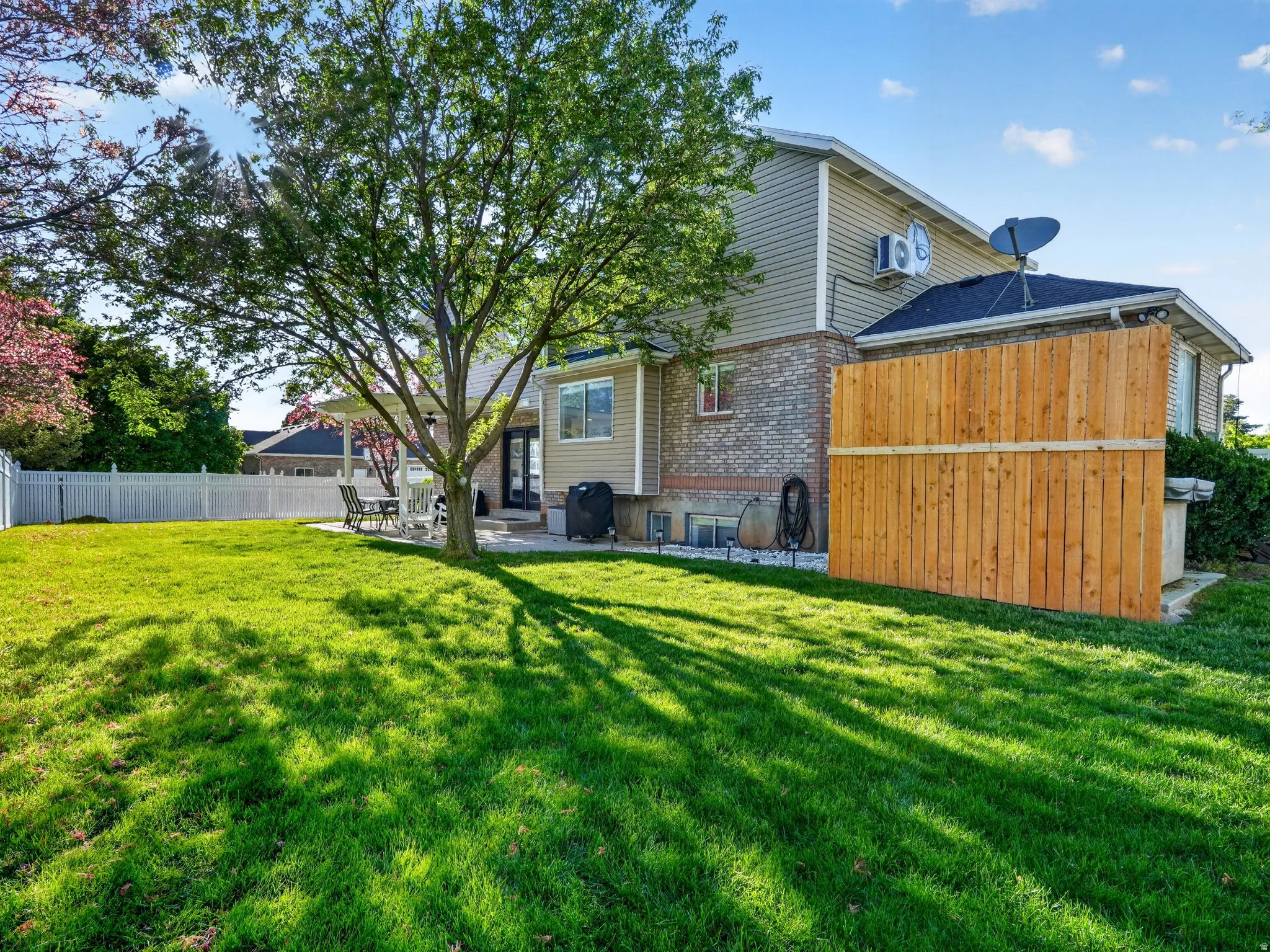 Expansive grassy yard featuring a prominent mature shade tree, a white picket fence, a wooden privacy fence, a brick and siding exterior, and a covered patio area